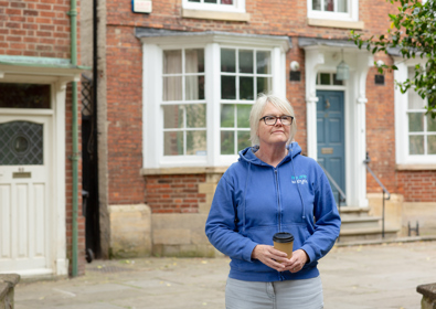 A middle-aged woman wearing glasses and a blue hoodie stands in front of a brick building holding a coffee cup. she appears thoughtful and content.
