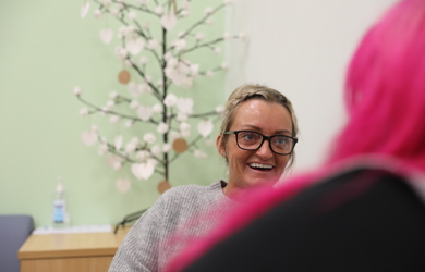 A woman with blonde hair and glasses smiling in a conversation with another person with pink hair. A decorative tree is in the background.