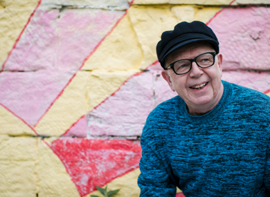 An smiling elderly man in glasses and a black cap. He sits before a colourfully painted wall.