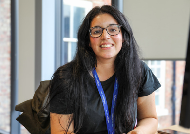 A smiling woman with dark hair and glasses, wearing a black shirt and a blue lanyard, sitting in an office with windows in the background.