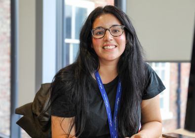 A smiling woman with dark hair and glasses, wearing a black shirt and a blue lanyard, sitting in an office with windows in the background.