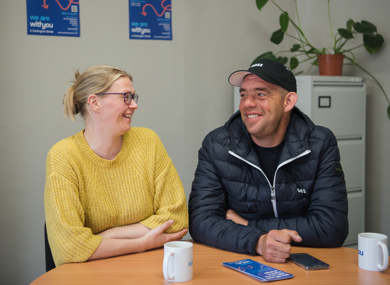 A man and a woman smiling at each other while sitting at a table with coffee mugs, pamphlets, and a plant in the background inside an office.