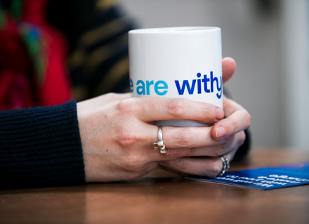 Close-up of a person's hands holding a white mug with the words "Withyou" in blue text.