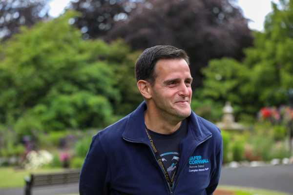A man in a "safer cornwall" uniform smiling outdoors with blurred greenery and trees in the background.