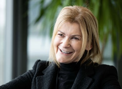 A woman wearing a black blazer and shirt, sits indoors by a window with a blurred cityscape background.
