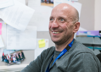 A smiling man sits at a desk in an office filled with papers and photographs, wearing a grey sweater and a blue lanyard.