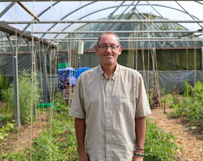 A middle-aged man smiling at the camera, standing in a greenhouse with various plants under a structure with transparent roofing.