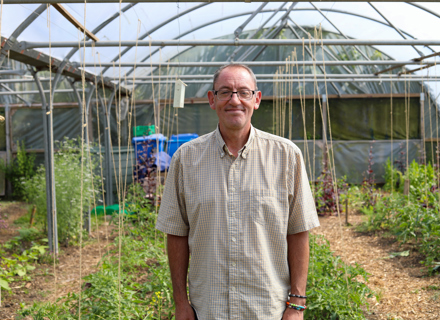 A middle-aged man smiling at the camera, standing in a greenhouse with various plants under a structure with transparent roofing.