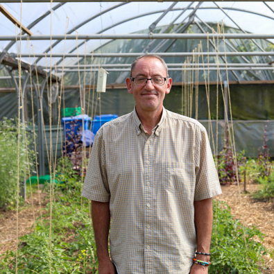 A middle-aged man smiling at the camera, standing in a greenhouse with various plants under a structure with transparent roofing.