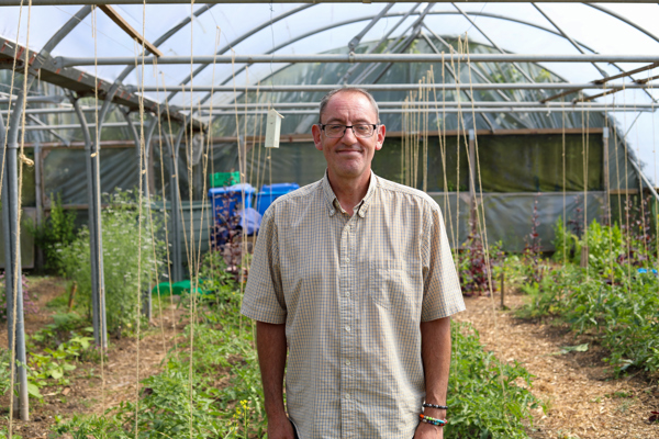 A middle-aged man smiling at the camera, standing in a greenhouse with various plants under a structure with transparent roofing.