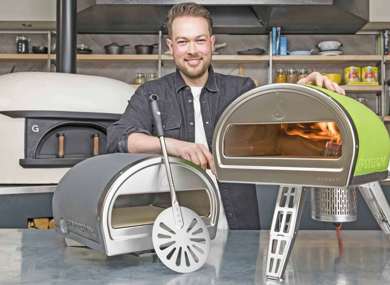A smiling man in a kitchen displays two portable pizza ovens on a counter, one open showing flames inside. He holds a pizza peel next to the ovens.