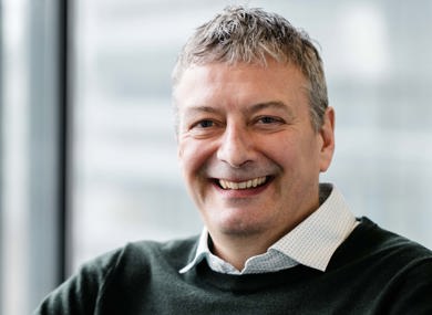 A middle-aged man , wearing a green sweater over a collared shirt, sits indoors by a window with a blurred cityscape background.