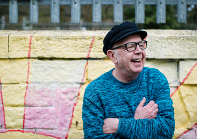 An smiling elderly man in glasses and a black cap. He sits before a colourfully painted wall.
