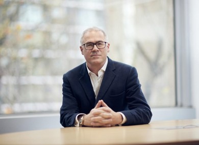 A middle-aged man in a dark suit sitting at a table, with hands clasped in front of him and a blurred background of windows showing trees.