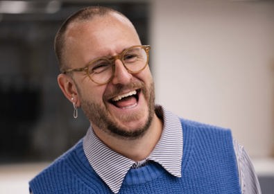 A man with a beard wearing glasses, a blue sweater vest, and a collared shirt, laughing in an office setting.