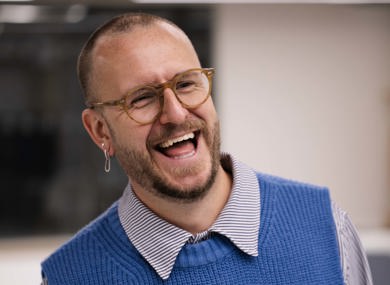 A man with a beard wearing glasses, a blue sweater vest, and a collared shirt, laughing in an office setting.
