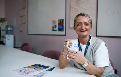 A smiling woman with a lanyard sits at a table holding a coffee mug, with informational pamphlets and a whiteboard visible in the background.