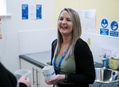 A woman smiling and holding a coffee mug in a kitchen with informational posters and sink in the background. she is conversing with another person.