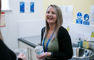 A woman smiling and holding a coffee mug in a kitchen with informational posters and sink in the background. she is conversing with another person.