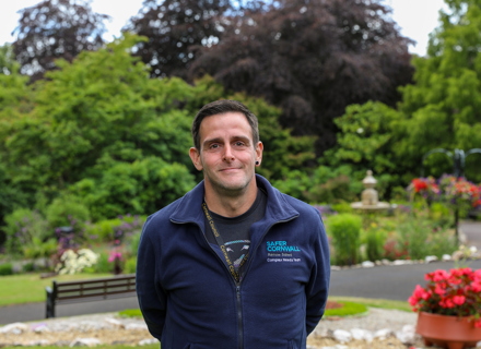 A man wearing a blue zip-up sweatshirt with "cornwall" embroidered on it is standing in a garden with trees, flowers, and a bench in the background.