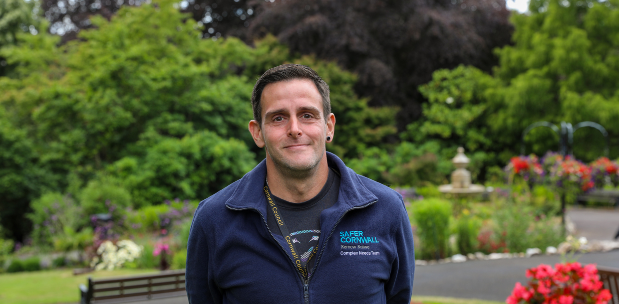 A man wearing a blue zip-up sweatshirt with "cornwall" embroidered on it is standing in a garden with trees, flowers, and a bench in the background.