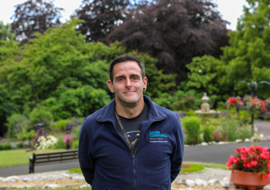 A man wearing a blue zip-up sweatshirt with "cornwall" embroidered on it is standing in a garden with trees, flowers, and a bench in the background.