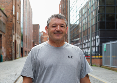 A smiling middle-aged man wearing a grey t-shirt stands in an urban alley with brick buildings and modern structures in the background.