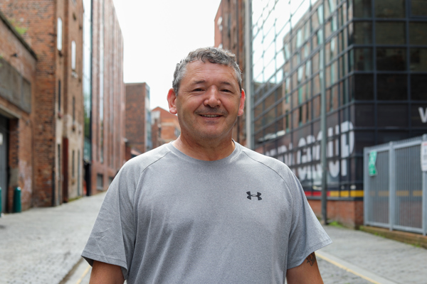 A smiling middle-aged man wearing a grey t-shirt stands in an urban alley with brick buildings and modern structures in the background.
