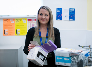 A smiling woman wearing a blue lanyard stands in an office holding medical supplies. notice boards with various flyers are visible behind her.