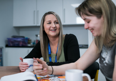 Two women sit at a kitchen table, smiling and chatting over coffee while looking at a magazine together. one woman has a blue lanyard around her neck.