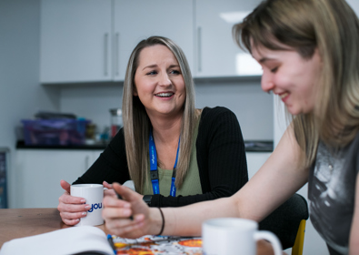 Two women sit at a kitchen table, smiling and chatting over coffee while looking at a magazine together. one woman has a blue lanyard around her neck.