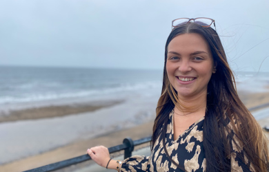 A smiling woman with sunglasses on her head, wearing a black and gold dress, stands by a railing with a beach and overcast sky in the background.
