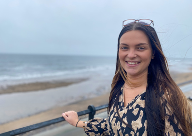 A smiling woman with sunglasses on her head, wearing a black and gold dress, stands by a railing with a beach and overcast sky in the background.