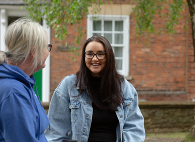 A young woman in glasses and a denim jacket smiles while engaging in a conversation with older woman, seen from the back, in front of a brick building.