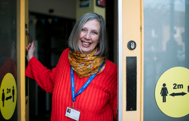 A cheerful woman with gray hair, wearing a red sweater and colorful scarf, stands at an open door smiling, with a lanyard and id around her neck.