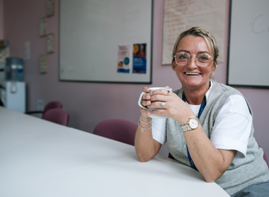 A smiling woman with a lanyard sits at a table holding a coffee mug, with informational pamphlets and a whiteboard visible in the background.