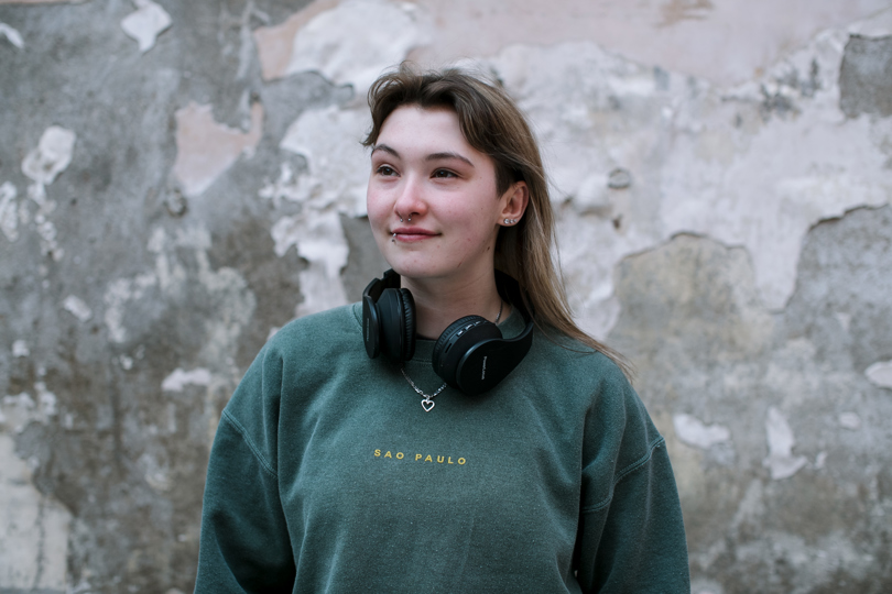 Chloe stands in front of a blank grey wall. She's wearing a dark green jumper and has black headphones around her neck. She looks slightly to the right and is smiling
