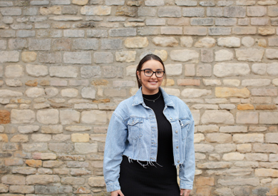 A smiling woman in a denim jacket and glasses stands in front of a textured stone wall.