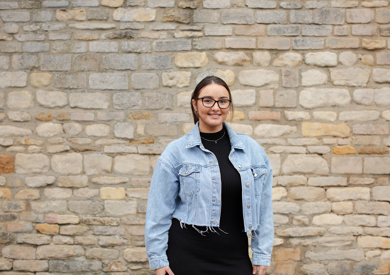 A smiling woman in a denim jacket and glasses stands in front of a textured stone wall.