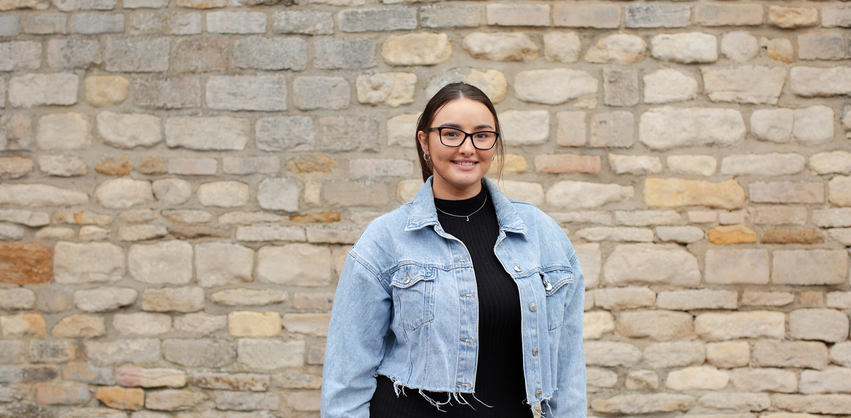 A smiling woman in a denim jacket and glasses stands in front of a textured stone wall.