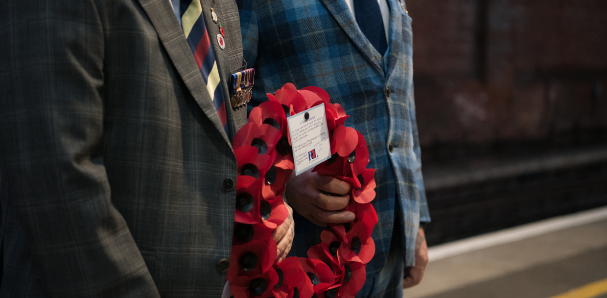 Two individuals in formal attire stand at a train station; one holds a wreath of red poppies and the other displays a medal and a ticket.