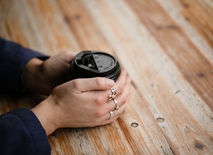 Close-up of a woman's hand holding a coffee cup on a wooden table, showing intricate rings on her fingers.