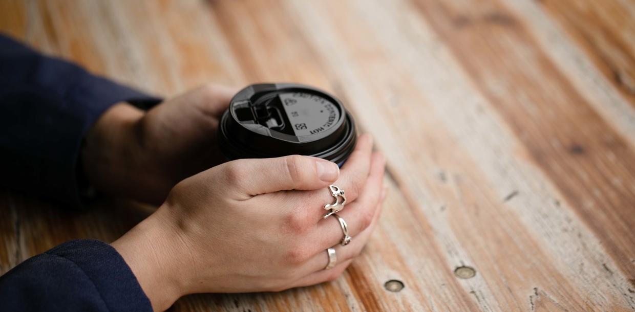 Close-up of a woman's hand holding a coffee cup on a wooden table, showing intricate rings on her fingers.