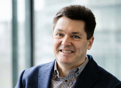 Portrait of a smiling man wearing a navy blazer and checkered shirt, sitting indoors with a window background that filters soft natural light.