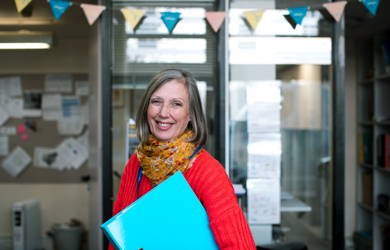 A smiling woman with grey hair, wearing a red sweater and a colourful scarf, holds a light blue folder in a room with glass doors in the background.