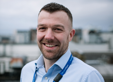 A man with short hair and a light beard, smiling at the camera. he wears a light blue shirt and a blue lanyard. Blurred cityscape under a cloudy sky in the background.