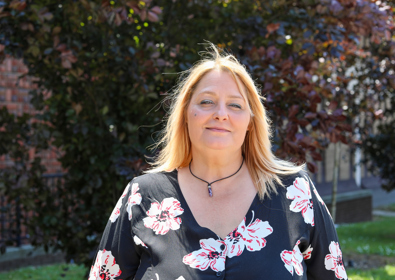 A woman with blonde hair, wearing a black blouse with red and white floral patterns, smiles gently. The background shows sunlit greenery.