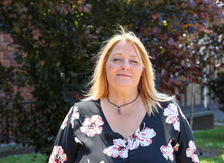 A woman with blonde hair, wearing a black blouse with red and white floral patterns, smiles gently. The background shows sunlit greenery.