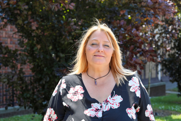 A woman with blonde hair, wearing a black blouse with red and white floral patterns, smiles gently. The background shows sunlit greenery.