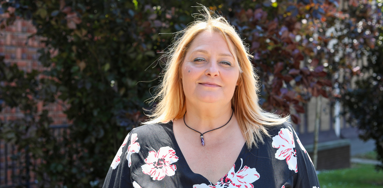 A woman with blonde hair, wearing a black blouse with red and white floral patterns, smiles gently. The background shows sunlit greenery.